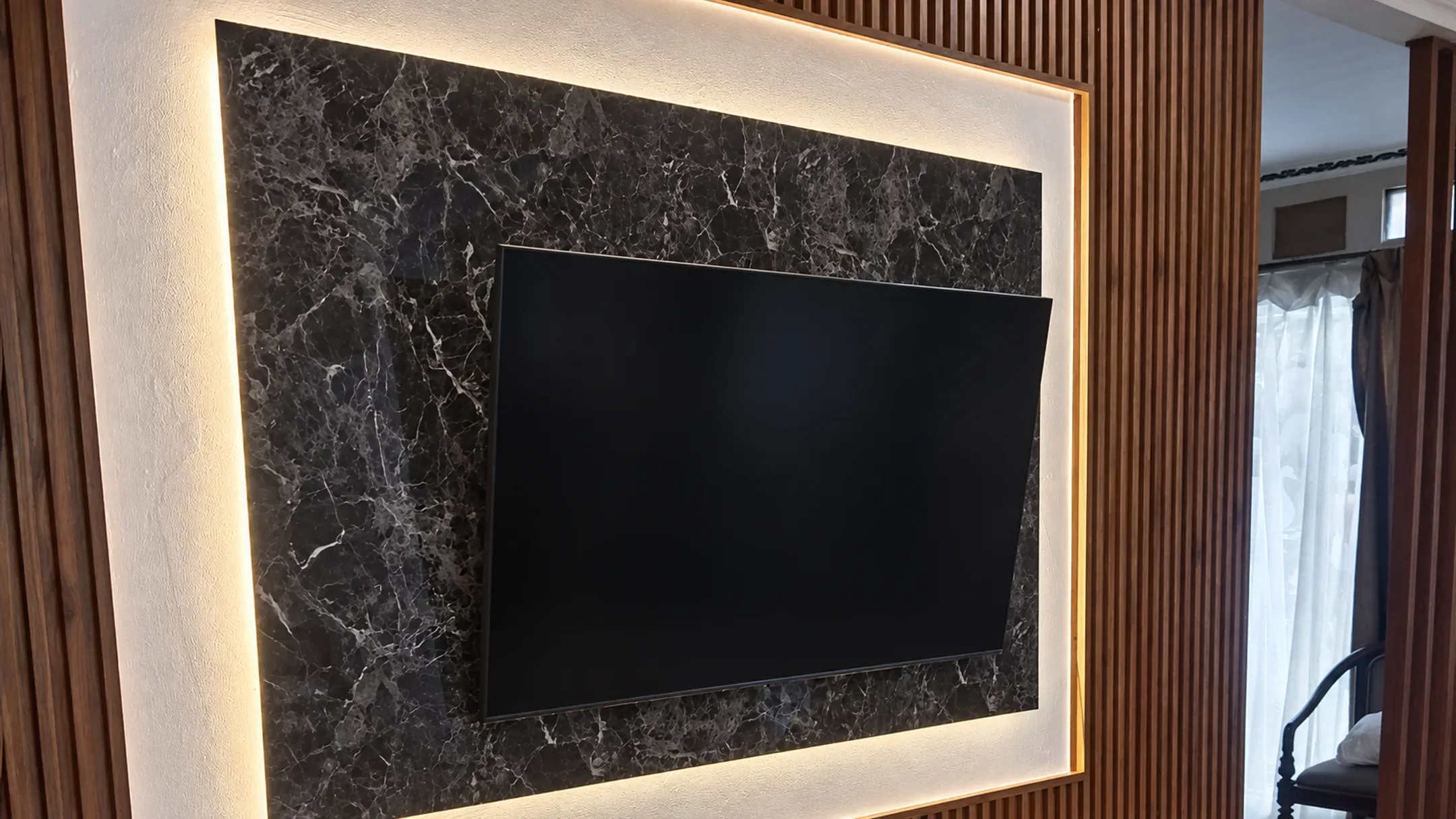 Foto pemasangan panel dinding motif kayu walnut gelap di sekeliling backdrop TV marmer hitam dengan lampu LED